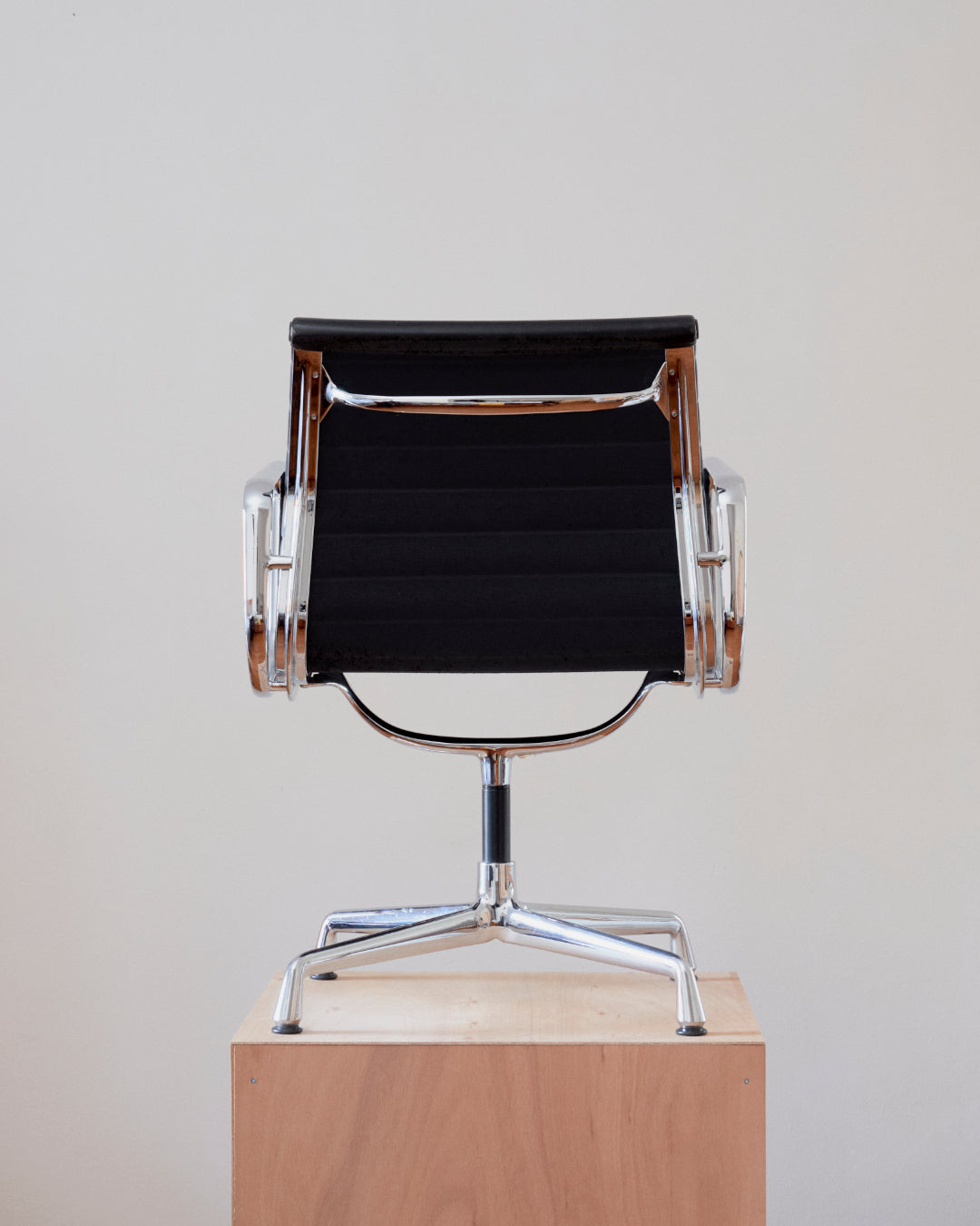 A modern armchair with a black fabric seat and backrest, supported by a polished chrome metal frame and star base, displayed on a light wood pedestal against a neutral off-white background.