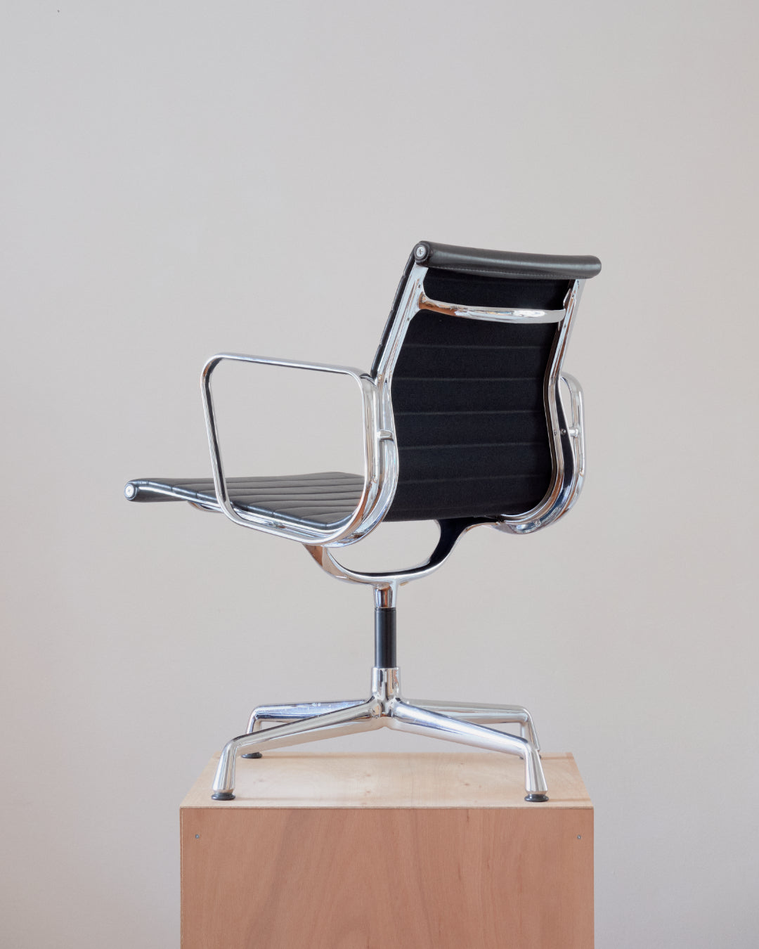 A modern black and chrome office chair with a ribbed mesh back and armrests, shown from the side on a light wood pedestal against a plain light gray background.