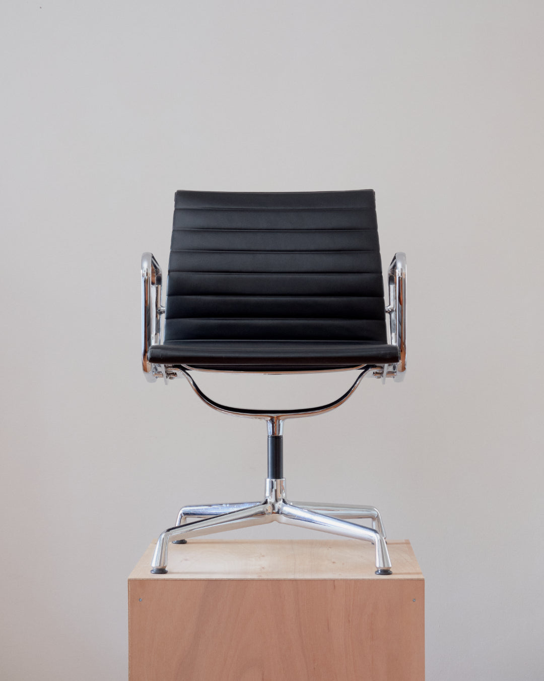 A modern black leather armchair with a ribbed high back and chrome metal base, displayed on a light wood pedestal against a plain light gray wall.