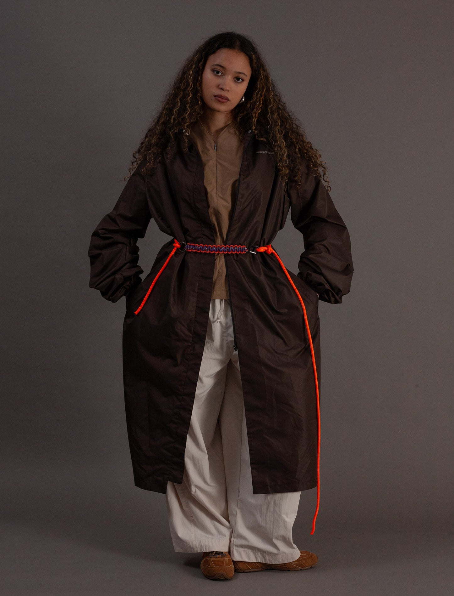 Person wearing a long brown coat with orange accents on a grey background
